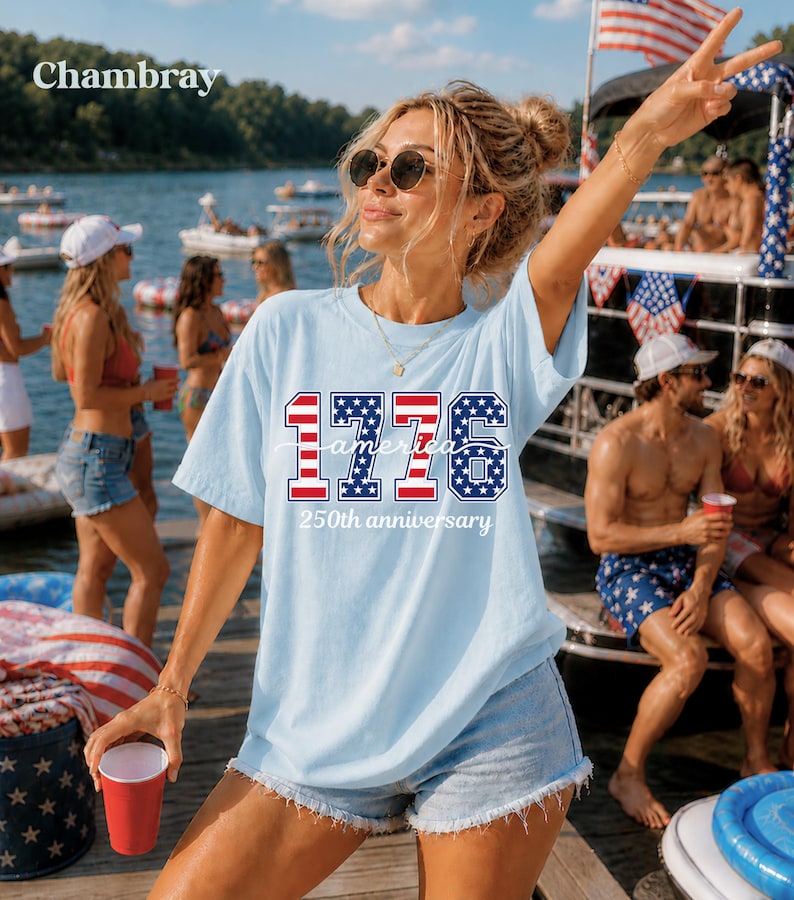 May include: A light blue t-shirt featuring the text "1776 America 250th anniversary" in red, white, and blue. The shirt is paired with denim shorts. The image is taken outdoors near a lake with people and boats in the background.