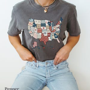 May include: A gray t-shirt with a patchwork design of the United States of America. The patches are red, white, and blue and feature words like "Brave", "Loved", "Free", and "Victorious".
