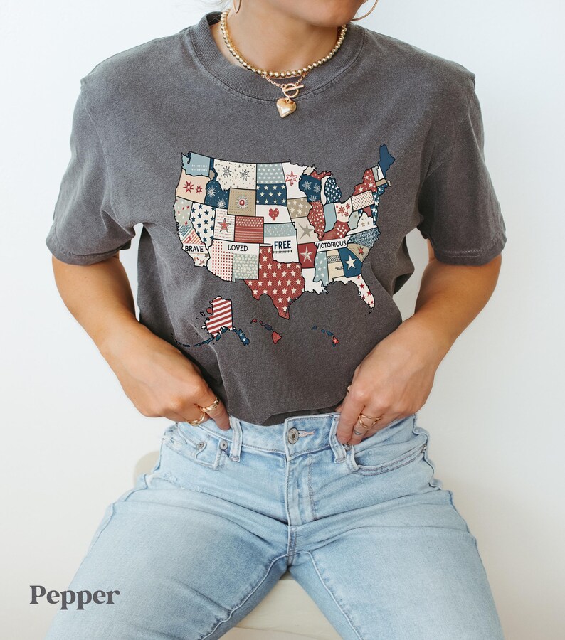 May include: A gray t-shirt with a patchwork design of the United States of America. The patches are red, white, and blue and feature words like "Brave", "Loved", "Free", and "Victorious".