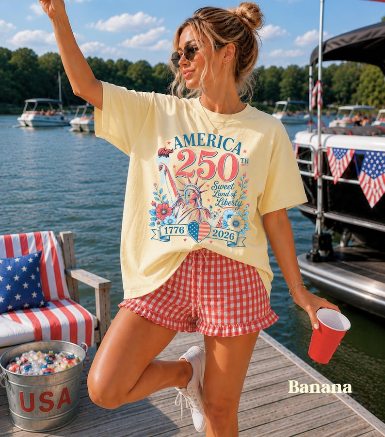K&ouml;nnte beinhalten: Hellgelbes T-Shirt mit einer Grafik zum 250. Jahrestag Amerikas, mit der Freiheitsstatue, Blumen und den Jahren 1776 und 2026. Kombiniert mit rot-wei&szlig; karierten Shorts.