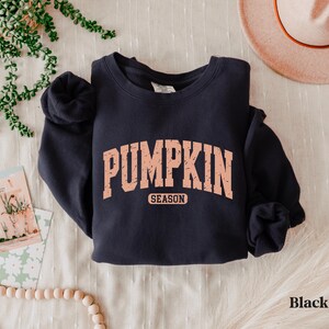 Puede incluir: Sudadera negra con el texto "PUMPKIN SEASON" en fuente color melocot&oacute;n estilo universitario. La sudadera est&aacute; doblada sobre una superficie blanca, con un sombrero y objetos decorativos en el fondo.