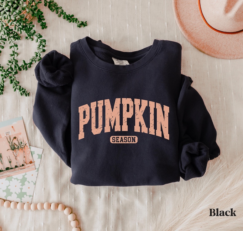 Puede incluir: Sudadera negra con el texto "PUMPKIN SEASON" en fuente color melocot&oacute;n estilo universitario. La sudadera est&aacute; doblada sobre una superficie blanca, con un sombrero y objetos decorativos en el fondo.