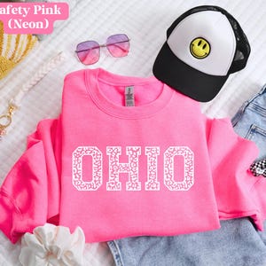 May include: A bright pink sweatshirt displaying the word "OHIO" in white, patterned letters. The image also features a black and white trucker cap with a yellow smiley face, pink sunglasses, and a keychain.