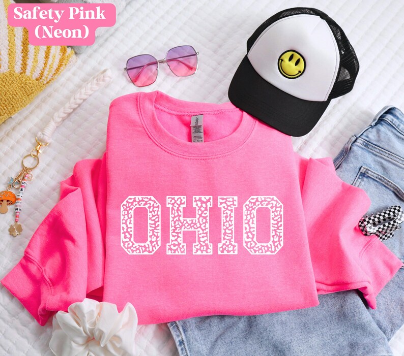 May include: A bright pink sweatshirt displaying the word "OHIO" in white, patterned letters. The image also features a black and white trucker cap with a yellow smiley face, pink sunglasses, and a keychain.