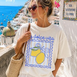 Pu&ograve; includere: T-shirt color avorio con un design "la dolce vita" con due limoni gialli e un nastro blu. La persona indossa occhiali da sole e una borsa di paglia. Lo sfondo mostra una citt&agrave; costiera con una chiesa e il mare.