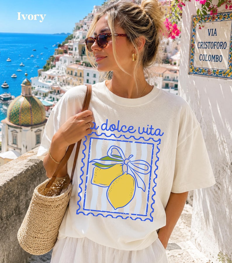 Pu&ograve; includere: T-shirt color avorio con un design "la dolce vita" con due limoni gialli e un nastro blu. La persona indossa occhiali da sole e una borsa di paglia. Lo sfondo mostra una citt&agrave; costiera con una chiesa e il mare.