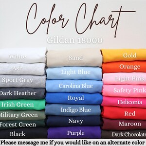 May include: A colour chart for Gildan 18000 sweatshirts featuring three vertical stacks of folded garments in various colours. Labels include White, Ash, Sport Gray, Dark Heather, Irish Green, Military Green, Forest Green, Black, Sand, Light Blue, Carolina Blue, Royal, Indigo Blue, Navy, Purple, Gold, Orange, Light Pink, Safety Pink, Heliconia, Red, Maroon, and Dark Chocolate.