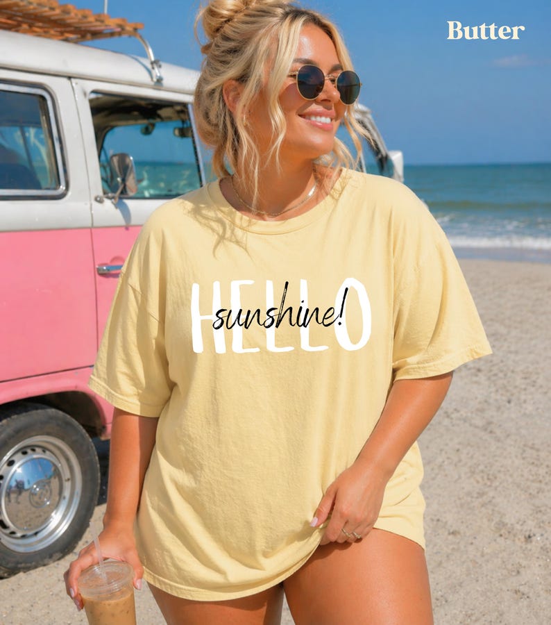 May include: A pale yellow t-shirt with the text "HELLO sunshine!" in white. The person is wearing sunglasses and is on a beach with a pink and white van in the background.