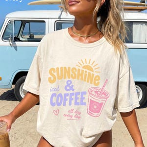 Puede incluir: Camiseta color marfil con las palabras "Sunshine iced & Coffee" en amarillo y morado, un gr&aacute;fico de taza de caf&eacute; rosa y la frase "all day every day". La camiseta se lleva delante de una furgoneta azul claro.