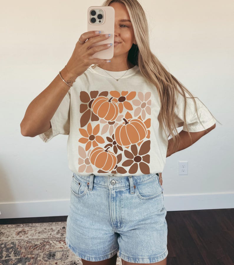 May include: Cream-colored t-shirt featuring a fall-themed design. The graphic includes orange pumpkins and flowers in shades of brown and beige. The shirt is paired with light blue denim shorts.