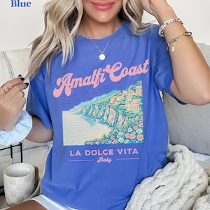 May include: A blue t-shirt with a graphic of the Amalfi Coast in Italy. The graphic includes a colorful illustration of the coastline, buildings, and flowers. The text "Amalfi Coast" is written in a pink and white font. The text "La Dolce Vita" is written in a black font below the graphic. The text "Italy" is written in a smaller black font below "La Dolce Vita".