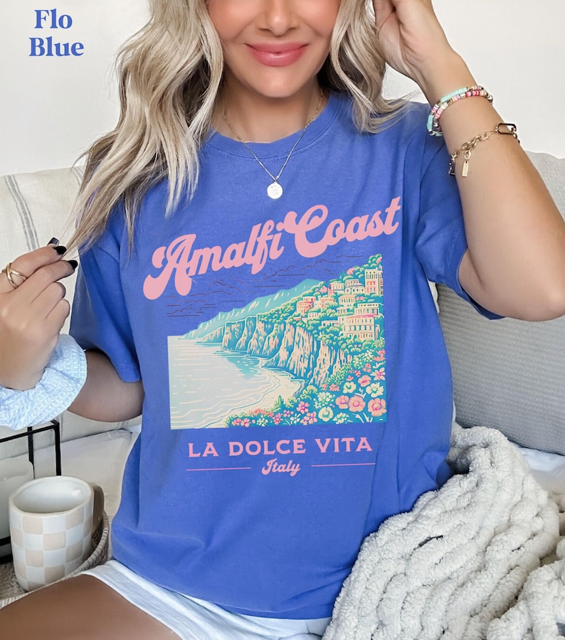 May include: A blue t-shirt with a graphic of the Amalfi Coast in Italy. The graphic includes a colorful illustration of the coastline, buildings, and flowers. The text "Amalfi Coast" is written in a pink and white font. The text "La Dolce Vita" is written in a black font below the graphic. The text "Italy" is written in a smaller black font below "La Dolce Vita".