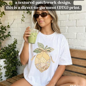 May include: White t-shirt with a direct-to-garment print of a lemon with a patchwork design. The lemon has a green leaf and bow, with yellow and floral patterned sections. The text on the shirt says "White".