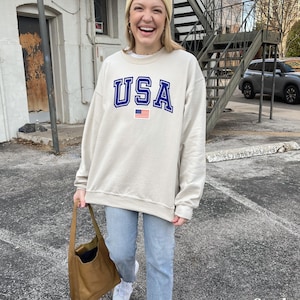 Pu&ograve; includere: Una felpa beige con una grafica USA blu e rossa e una piccola bandiera americana. La felpa &egrave; indossata da una persona che indossa jeans blu e un berretto beige.