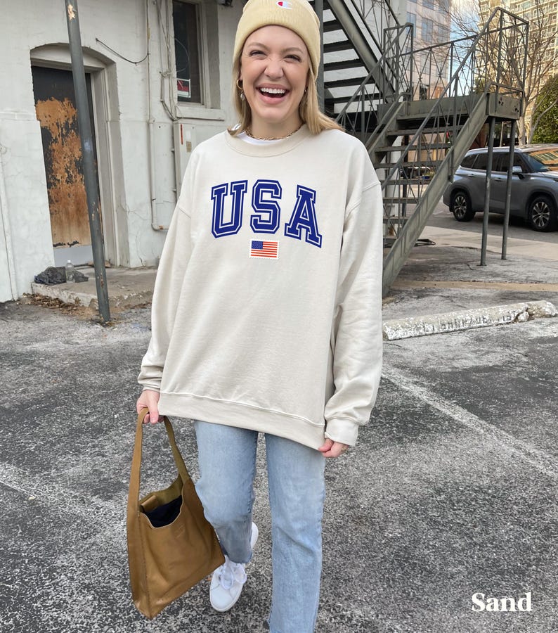 Pu&ograve; includere: Una felpa beige con una grafica USA blu e rossa e una piccola bandiera americana. La felpa &egrave; indossata da una persona che indossa jeans blu e un berretto beige.