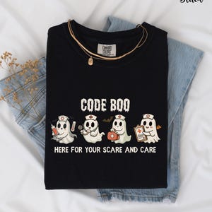 May include: Black t-shirt with the text "CODE BOO" and four cartoon ghost characters dressed as medical professionals. The ghosts hold a syringe, stethoscope, first aid kit, and clipboard. The phrase "HERE FOR YOUR SCARE AND CARE" is below the ghosts.