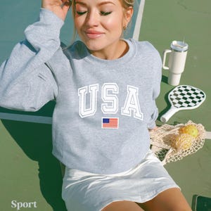 Pu&ograve; includere: Una felpa grigia con la scritta "USA" stampata in bianco con una piccola bandiera americana sotto. La felpa &egrave; indossata da una persona su un campo da tennis.