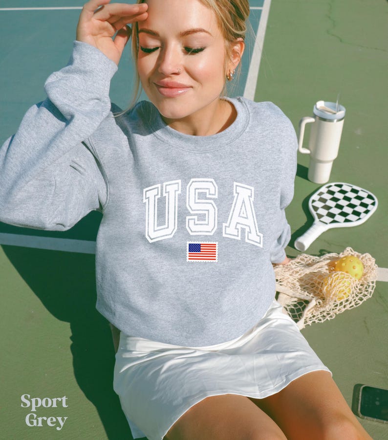 Pu&ograve; includere: Una felpa grigia con la scritta "USA" stampata in bianco con una piccola bandiera americana sotto. La felpa &egrave; indossata da una persona su un campo da tennis.