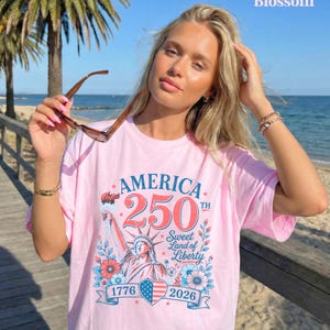 K&ouml;nnte beinhalten: Hellrosa T-Shirt mit dem Text "AMERICA 250TH Sweet Land of Liberty 1776-2026" und einer Grafik der Freiheitsstatue. Die Person tr&auml;gt eine Sonnenbrille.