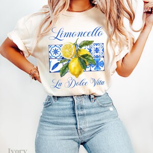 Pu&ograve; includere: Una maglietta bianca con il testo "Limoncello La Dolce Vita" e una grafica di limoni e piastrelle blu e bianche.