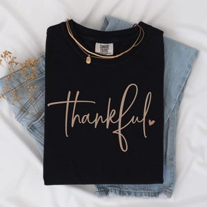 May include: Black t-shirt with the word "thankful" in a cursive font and a small heart. The shirt is folded on top of blue jeans and a gold necklace. The word "Black" is in the top right corner.