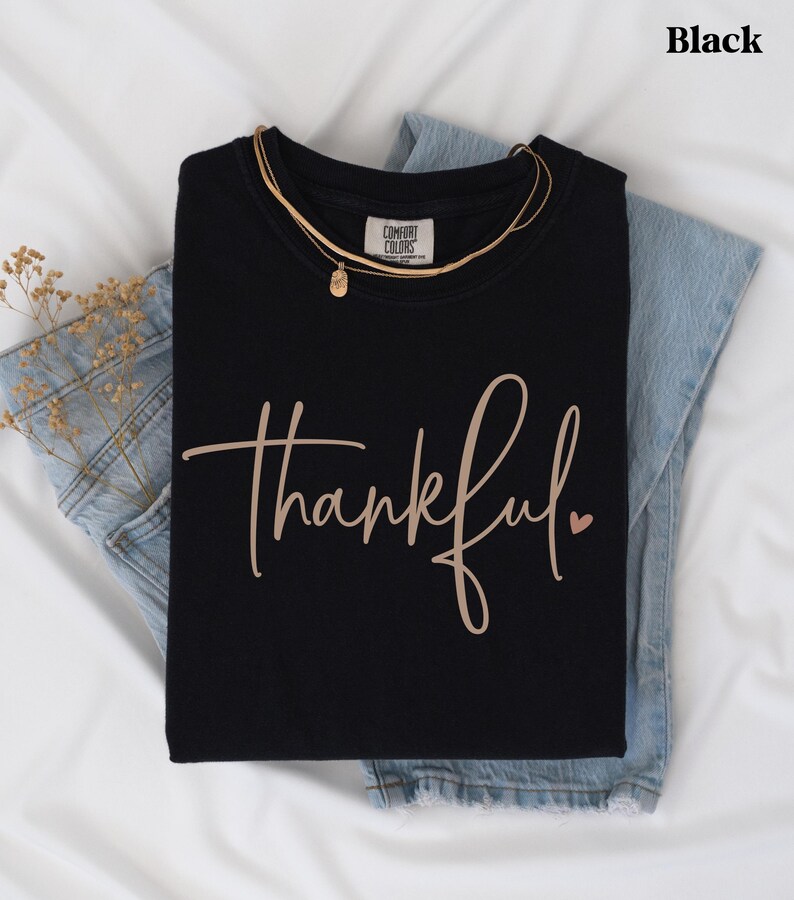 May include: Black t-shirt with the word "thankful" in a cursive font and a small heart. The shirt is folded on top of blue jeans and a gold necklace. The word "Black" is in the top right corner.