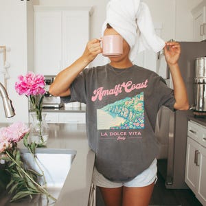 May include: A woman wearing a gray t-shirt with a graphic of the Amalfi Coast and the text "Amalfi Coast, La Dolce Vita, Italy". She is holding a pink mug and has a white towel wrapped around her head.