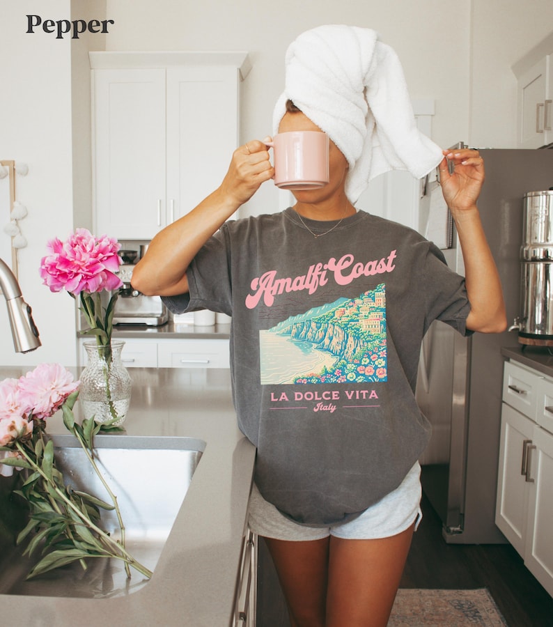 May include: A woman wearing a gray t-shirt with a graphic of the Amalfi Coast and the text "Amalfi Coast, La Dolce Vita, Italy". She is holding a pink mug and has a white towel wrapped around her head.