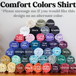 May include: A display of rolled Comfort Colors shirts in various colors, including white, black, and blue. The text at the top reads "Comfort Colors Shirt." Additional text encourages inquiries about alternate colors.