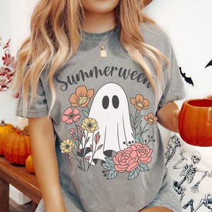 May include: Grey t-shirt with the word "Summerween" above a white ghost illustration surrounded by colorful flowers. The ghost has two black oval eyes. The flowers are in shades of pink, yellow, and orange.