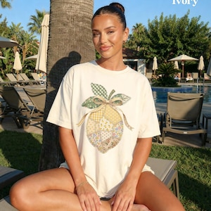 May include: Ivory-coloured t-shirt featuring a patchwork lemon design with green, yellow, and floral patterns. The shirt has a relaxed fit and short sleeves. The word "Ivory" is printed in white in the upper right corner.