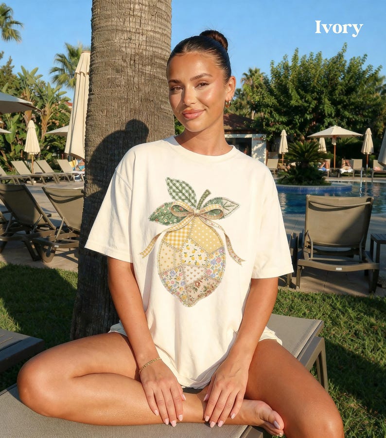 May include: Ivory-coloured t-shirt featuring a patchwork lemon design with green, yellow, and floral patterns. The shirt has a relaxed fit and short sleeves. The word "Ivory" is printed in white in the upper right corner.