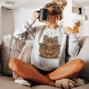 Op de afbeelding: Een sportgrijze sweater met lange mouwen en een afbeelding van een oranje pompoen met een zonnebril met luipaardprint en een bijpassende strik. De tekst Sport Grey staat in de linkerbovenhoek van de afbeelding.