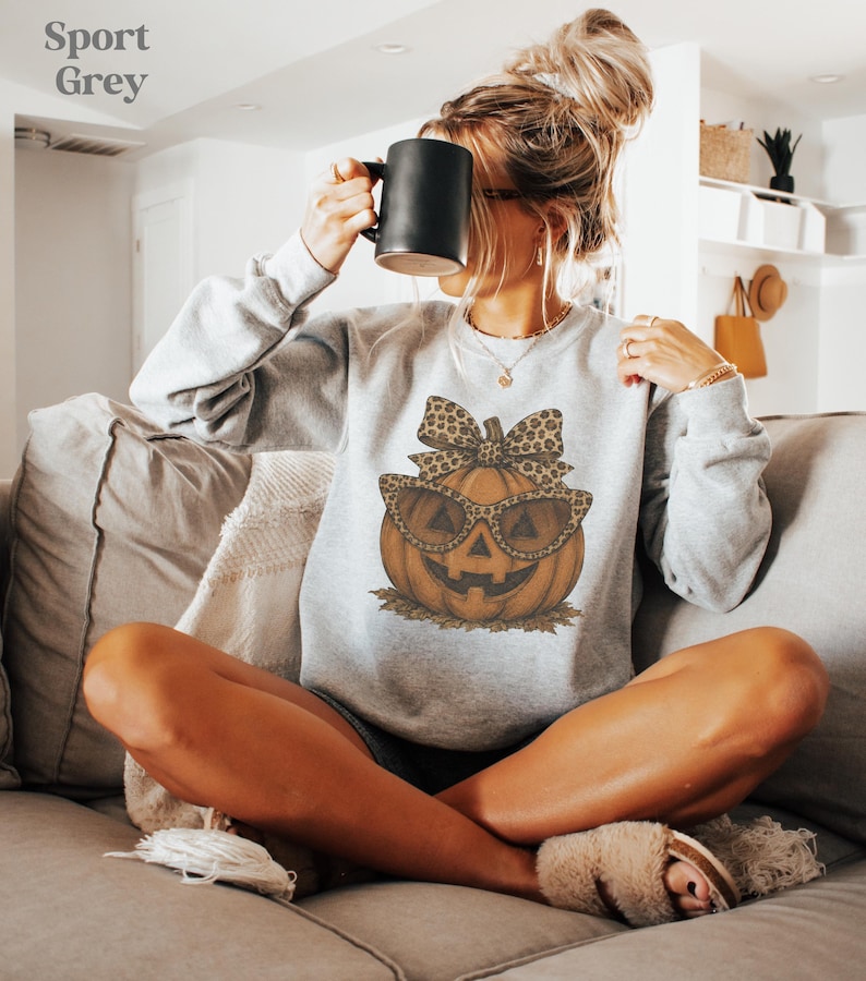 Op de afbeelding: Een sportgrijze sweater met lange mouwen en een afbeelding van een oranje pompoen met een zonnebril met luipaardprint en een bijpassende strik. De tekst Sport Grey staat in de linkerbovenhoek van de afbeelding.