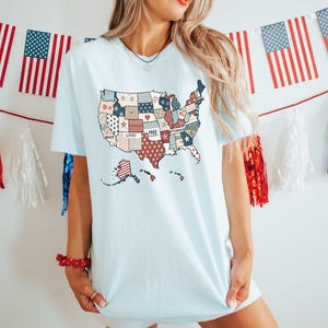 May include: A light blue t-shirt with a patchwork design of the United States. The patches are red, white, and blue and feature words like "Brave", "Loved", "Free", and "Victorious".