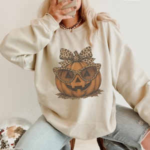 Op de afbeelding: Een zandkleurige sweater met ronde hals en een grafische print van een pompoen met een zonnebril met luipaardprint en een bijpassende strik. De tekst "Sand" staat in de rechterbovenhoek van de afbeelding gedrukt.
