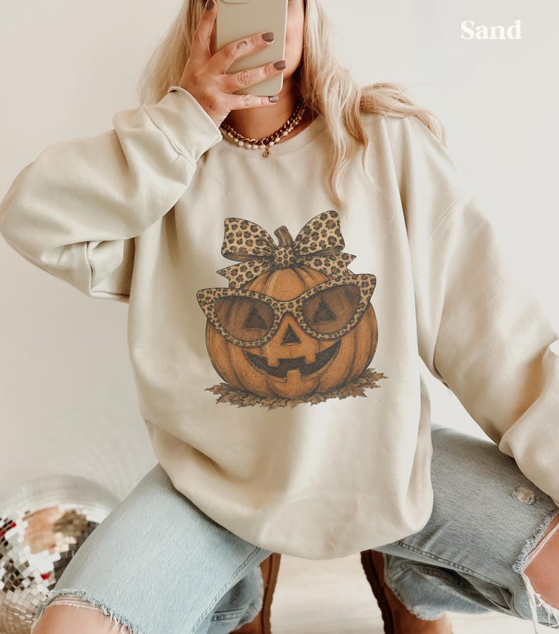 Op de afbeelding: Een zandkleurige sweater met ronde hals en een grafische print van een pompoen met een zonnebril met luipaardprint en een bijpassende strik. De tekst "Sand" staat in de rechterbovenhoek van de afbeelding gedrukt.