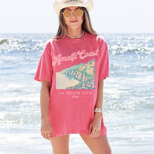 May include: A pink oversized t-shirt with a graphic print of a coastal scene and the text "Amalfi Coast" in a retro font. The text "La Dolce Vita" and "Italy" are also printed on the shirt.