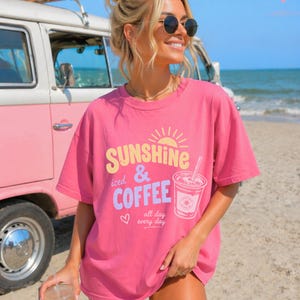Puede incluir: Una camiseta rosa con la frase "Sunshine & Iced Coffee all day every day" y un gr&aacute;fico de una taza de caf&eacute;. La persona lleva la camiseta en una playa, con una furgoneta rosa y blanca al fondo.