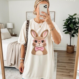 May include: Ivory-colored t-shirt featuring a detailed graphic of a bunny wearing pink glasses and a bow tie. The bunny has brown and pink accents. The shirt is a relaxed fit, suitable for casual wear.