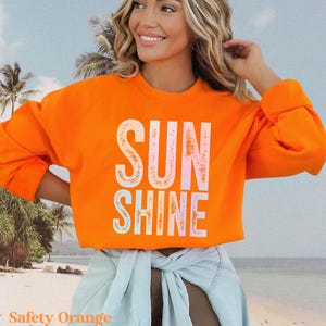 May include: Safety orange neon sweatshirt with the word "SUNSHINE" in a white, light pink, and light blue font. The sweatshirt has long sleeves and a crew neck. The model is wearing a light blue denim jacket tied around the waist.