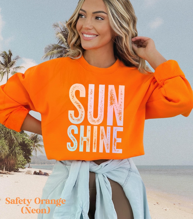 May include: Safety orange neon sweatshirt with the word "SUNSHINE" in a white, light pink, and light blue font. The sweatshirt has long sleeves and a crew neck. The model is wearing a light blue denim jacket tied around the waist.