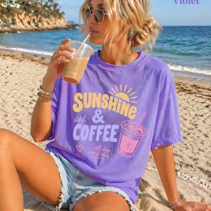 Puede incluir: Una camiseta violeta con las palabras "SUNSHINE iced & COFFEE" en amarillo y rosa, con un dise&ntilde;o de sol y taza de caf&eacute;. La camiseta se usa con pantalones cortos de mezclilla en una playa.