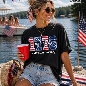 May include: Black t-shirt featuring the text "1776 America 250th anniversary" in red, white, and blue. The shirt is paired with denim shorts. A red cup and a straw bag are also visible.
