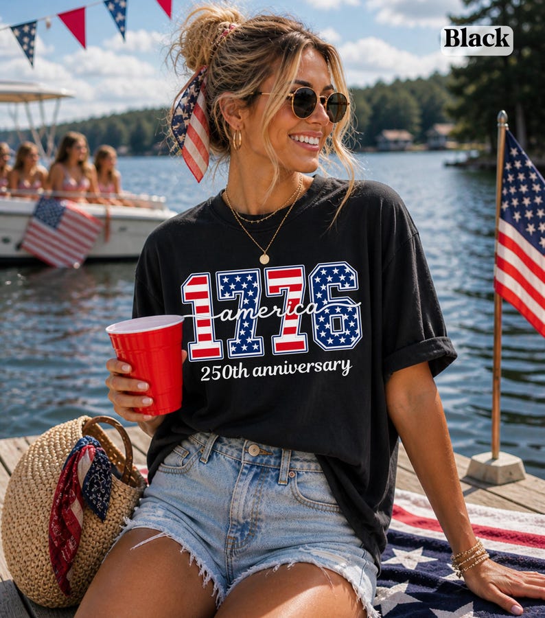 May include: Black t-shirt featuring the text "1776 America 250th anniversary" in red, white, and blue. The shirt is paired with denim shorts. A red cup and a straw bag are also visible.
