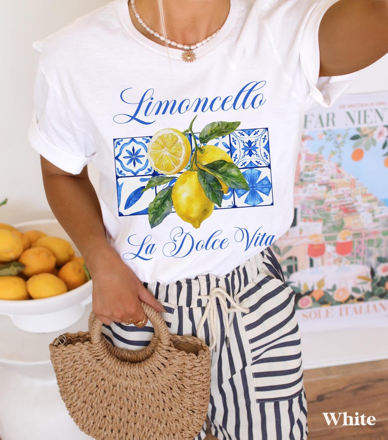 Pu&ograve; includere: Maglietta bianca con una grafica di limoni e piastrelle blu e bianche. Il testo "Limoncello La Dolce Vita" &egrave; stampato sulla maglietta.
