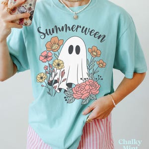 May include: A light blue t-shirt with the word "Summerween" in black script above a white ghost illustration surrounded by colorful flowers. The shirt has a relaxed fit and short sleeves. The text "Chalky Mint" is in the bottom right corner.
