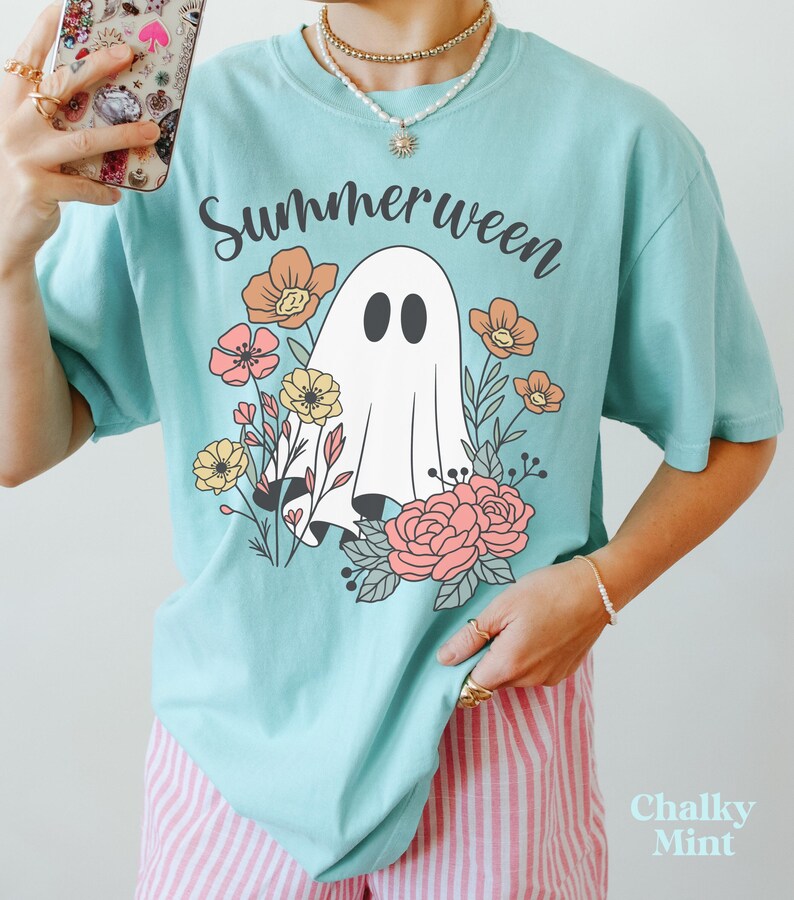 May include: A light blue t-shirt with the word "Summerween" in black script above a white ghost illustration surrounded by colorful flowers. The shirt has a relaxed fit and short sleeves. The text "Chalky Mint" is in the bottom right corner.