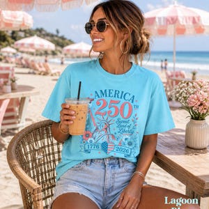 K&ouml;nnte beinhalten: Ein lagunenblaues T-Shirt mit dem Text "America 250th Sweet Land of Liberty 1776-2026" in Rot, Wei&szlig; und Blau. Das Shirt zeigt eine Grafik der Freiheitsstatue und florale Akzente. Das Model tr&auml;gt Jeansshorts und eine Sonnenbrille.