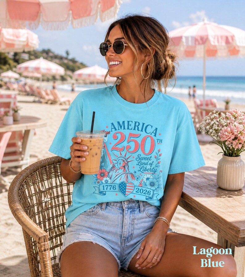 K&ouml;nnte beinhalten: Ein lagunenblaues T-Shirt mit dem Text "America 250th Sweet Land of Liberty 1776-2026" in Rot, Wei&szlig; und Blau. Das Shirt zeigt eine Grafik der Freiheitsstatue und florale Akzente. Das Model tr&auml;gt Jeansshorts und eine Sonnenbrille.